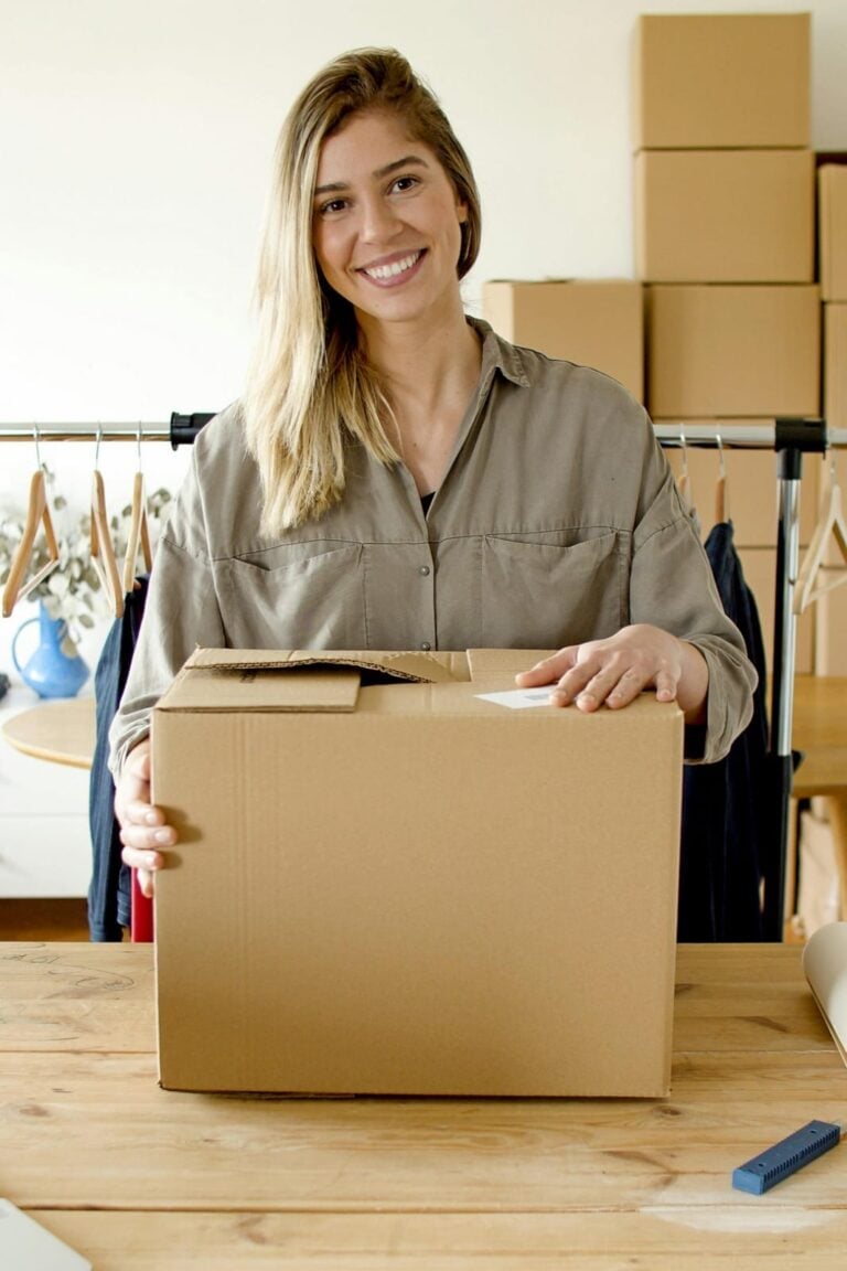 Woman in beige shirt smiling while holding an open cardboard box at a wooden table with stacked boxes in background
